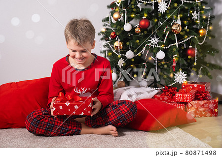 Happy child with blonde hair is sitting on floor at home and holding Christmas present near decorated Christmas tree Happy child with blonde hair is sitting on floor at home and holding Christmas present near decorated Christmas tree 80871498