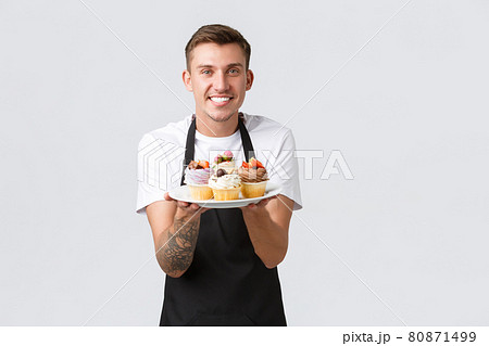 Small retail business, coffee shops and bakery concept. Friendly-looking handsome barista showing new menu, delicious cupcakes on plate, waiter serving order, tasty muffins over white background Small retail business, coffee shops and bakery concept. Friendly-looking handsome barista showing new menu, delicious cupcakes on plate, waiter serving order, tasty muffins over white background 80871499