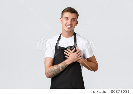 Small business owners, coffee shop and staff concept. Happy handsome blond guy in black apron, barista or waiter holding hand on heart and smiling, cherish cafe guests, love every customer 80873051
