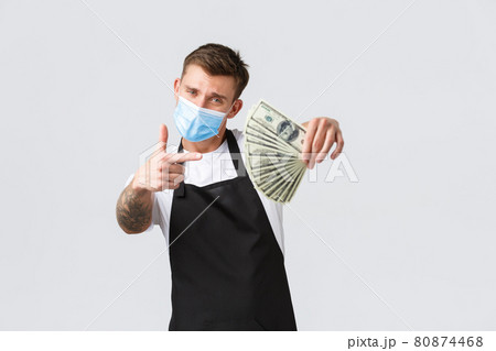 Small retail business, employees and coffee shop concept. Sassy good-looking barista, cafe owner in medical mask and apron, showing money, make thumbs-up gesture, suggest job with good payment Small retail business, employees and coffee shop concept. Sassy good-looking barista, cafe owner in medical mask and apron, showing money, make thumbs-up gesture, suggest job with good payment 80874468