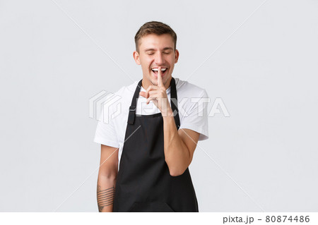 Small business owners, coffee shop and staff concept. Handsome happy barista, waiter in cafe wearing black apron, laughing and make shush gesture, asking keep quiet or not tell secret 80874486