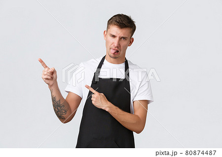 Small business owners, coffee shop and staff concept. Disappointed grimacing barista in black apron, pointing upper left corner, showing tongue from disgust or dislike, standing white background 80874487