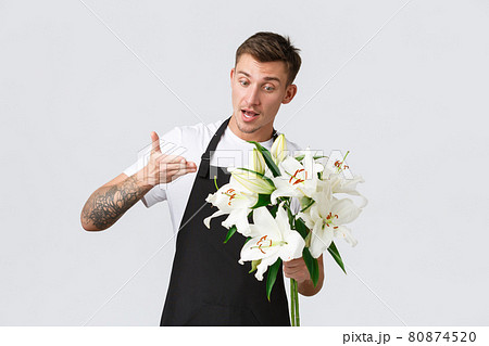 Small business, retail and employees concept. Charismatic handsome florist, salesman in flower shop selling bouquet, pointing at lilies, talking to client, standing white background Small business, retail and employees concept. Charismatic handsome florist, salesman in flower shop selling bouquet, pointing at lilies, talking to client, standing white background 80874520