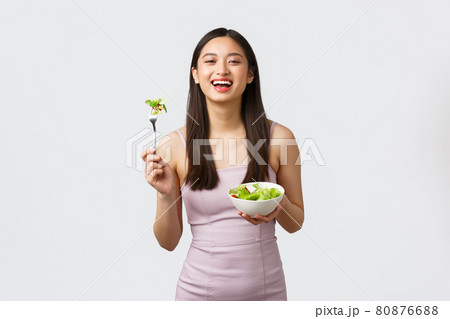 Healthy lifestyle, leisure and people emotions concept. Portrait of happy asian girl eating salad in beautiful evening dress, have bite of fresh vegetables, standing white background Healthy lifestyle, leisure and people emotions concept. Portrait of happy asian girl eating salad in beautiful evening dress, have bite of fresh vegetables, standing white background 80876688