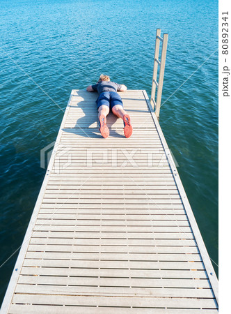 Boy lying on wooden pier over a lake watch fish in  blue water below 80892341