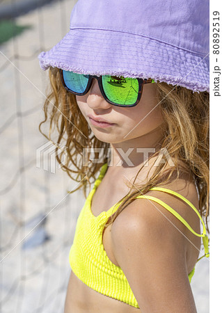 Portrait of a young beautiful model girl with hat and sunglasses posing on the beach. Wearing a bright yellow bikini swimsuit. Summer day, white sand.  80892519