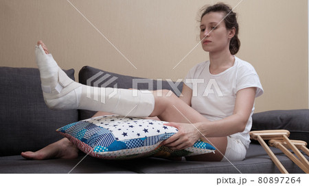 woman with a plaster cast sits on a sofa, a pillow is placed under a broken leg. woman with a plaster cast sits on a sofa, a pillow is placed under a broken leg. 80897264