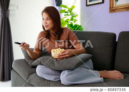 Mid adult Asian woman sitting on sofa in home living room holding bolw of popcorn and using remote control aiming to tv while watching in relax time 80897336