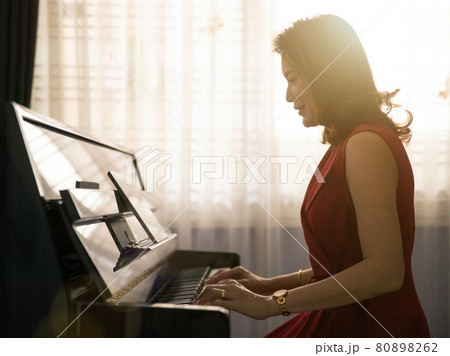 Middle age dignified Asian woman learning and studying new skills how to play the piano via online class from a tablet computer with happiness. Beautiful orange sunlight comes from window in the back 80898262