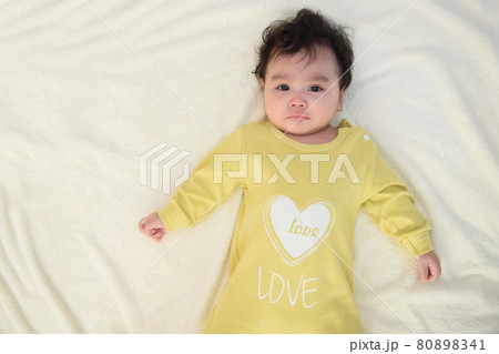 A cute little Asian baby in a yellow dress is lying on the white bed looking at the camera making a sad expression A cute little Asian baby in a yellow dress is lying on the white bed looking at the camera making a sad expression 80898341