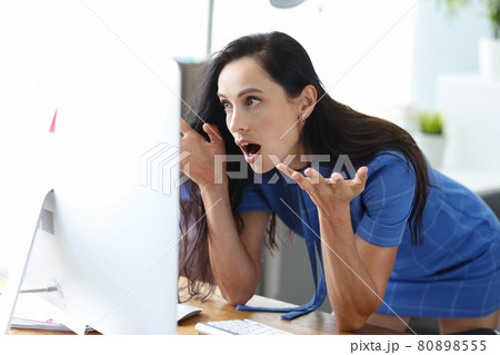 Shocked woman sits at table and looks at computer screen eyes widening in horror Shocked woman sits at table and looks at computer screen eyes widening in horror 80898555
