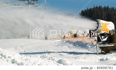 Snow making system. Snow cannon makes artificial snow of water on sunny day in ski resort. On backdrop skiers slide down snow-covered white slope of mountain, ski lift working, winter forest, blue sky Snow making system. Snow cannon makes artificial snow of water on sunny day in ski resort. On backdrop skiers slide down snow-covered white slope of mountain, ski lift working, winter forest, blue sky 80898781