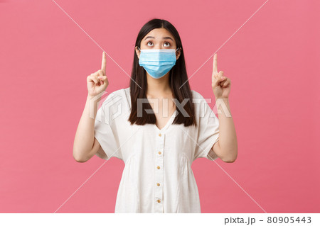 Covid-19, social distancing, virus and lifestyle concept. Curious pretty asian girl in medical mask and summer dress, looking and pointing fingers up at promo, advertisement, pink background 80905443