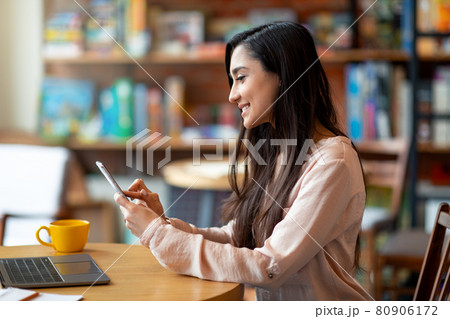 Happy latin woman holding mobile phone and using laptop, freelancer working at cafe, copy space, side view 80906172