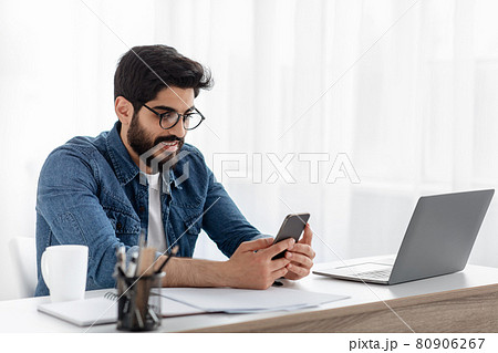 Arab freelancer guy using cellphone while working on laptop at desk at home office, browsing new app or messaging 80906267