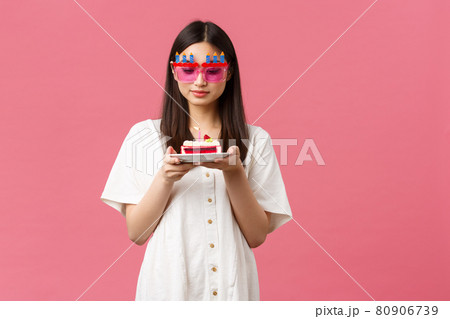 Celebration, party holidays and fun concept. Dreamy cute birthday girl in funny sunglasses holding b-day cake and gazing at candle thoughtful, making wish, standing pink background 80906739