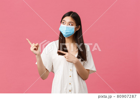 Covid-19, social distancing, virus and lifestyle concept. Curious cute asian girl, store visitor in medical mask asking question about product, pointing fingers left, standing pink background 80907287