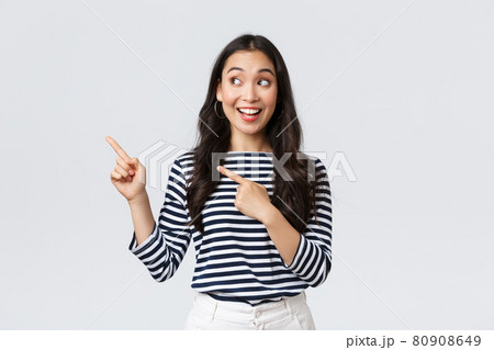 Lifestyle, people emotions concept. Excited good-looking asian girl smiling pleased as found excellent product, pointing fingers left at advertisement and looking satisfied, recommend promo 80908649