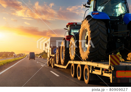POV heavy industrial truck semi trailer flatbed platform transport two big modern farming tractor machine on common highway road at sunset sunrise sky. Agricultural equipment transportation service 80909027
