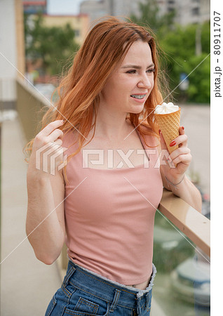 Portrait of young beautiful red-haired woman smiling with braces and going to eat ice cream cone outdoors in summer Portrait of young beautiful red-haired woman smiling with braces and going to eat ice cream cone outdoors in summer 80911707
