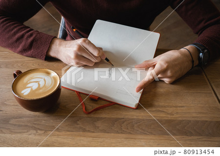 A young man sits in cafe, drinks coffee and makes notes and sketches in notebook. A young man sits in cafe, drinks coffee and makes notes and sketches in notebook. 80913454