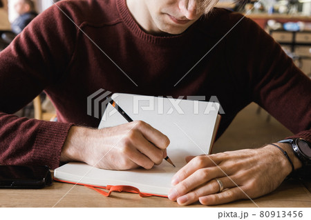 A young man sits in cafe, drinks coffee and makes notes and sketches in notebook. A young man sits in cafe, drinks coffee and makes notes and sketches in notebook. 80913456