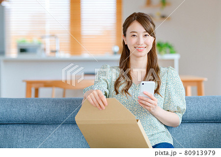 リビングでメール便の荷物を開けるスマホを持った若い主婦 80913979