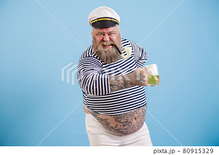 Happy plus size man in sailor suit holds delicious beer on light blue background Happy plus size man in sailor suit holds delicious beer on light blue background 80915329