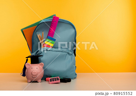 Blue backpack with school tools and piggy bank in hat on yellow background Blue backpack with school tools and piggy bank in hat on yellow background 80915552