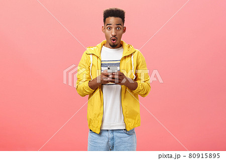 Impressed enthusiastic african american male with curly haircut folding lips in wow sound holding smartphone being excited and thrilled with cool device feautres or new app over pink wall 80915895