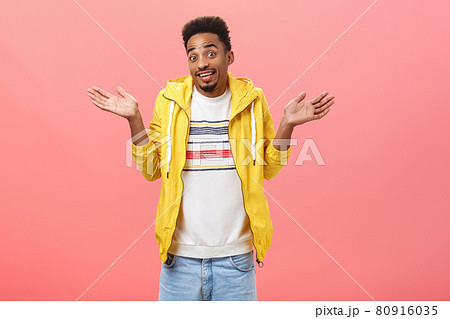 Have no idea what you talking about. Portrait of carefree unaware and clueless charming african american guy in yellow trendy jacket making shoulder shrug with spread hands being unsure over pink wall 80916035