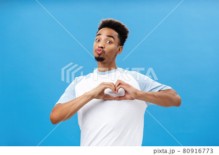 Funny and cute dark-skinned young male student in t-shirt folding lips in mwah or kiss showing heart gesture over breast giving love and affection signs to girl he likes over blue wall 80916773