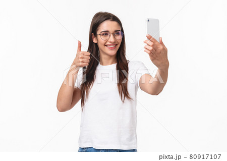 Communication, technology and lifestyle concept. Portrait of upbeat happy, smiling caucasian girl in glasses, internet blogger taking selfie, record video message to followers thumbs-up, hold phone Communication, technology and lifestyle concept. Portrait of upbeat happy, smiling caucasian girl in glasses, internet blogger taking selfie, record video message to followers thumbs-up, hold phone 80917107