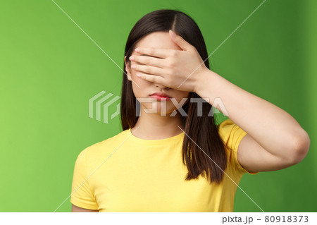 Sad tired asian girl dark haircut, shut eyes hold hand sight, grimacing, feeling gloomy and upset, stand green background depressed, unwilling watch how life ruins, pose yellow t-shirt 80918373