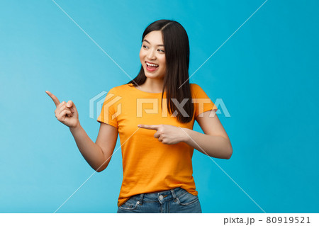 Friendly enthusiastic asian girl curiously looking sideways, pointing left interesting object, grinning joyfully discuss new opened store, stand blue background carefree lively talking 80919521