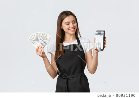 Grocery store employees, small business and coffee shops concept. Smiling pleased barista in black apron looking at payment terminal satisfied as showing cash, prefer POS paying 80921190