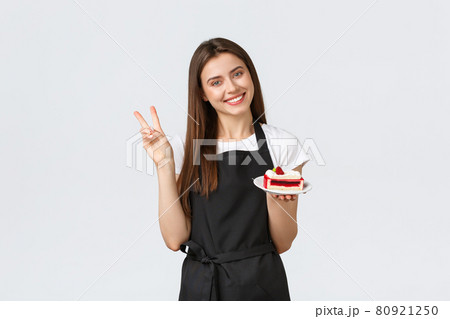 Grocery store employees, small business and coffee shops concept. Friendly smiling cute barista in black apron showing peace sign as serving delicious cake, working in cafe, white background Grocery store employees, small business and coffee shops concept. Friendly smiling cute barista in black apron showing peace sign as serving delicious cake, working in cafe, white background 80921250