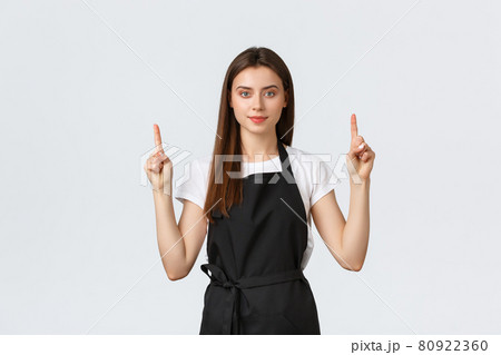 Small business, employees and coffee shop concept. Confient pretty barista in black apron pointing fingers up. Cafe employee showing advertisement. Cheerful saleswoman recommend special promo Small business, employees and coffee shop concept. Confient pretty barista in black apron pointing fingers up. Cafe employee showing advertisement. Cheerful saleswoman recommend special promo 80922360