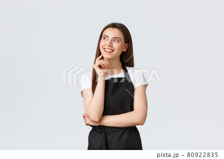 Small business, employees and coffee shop concept. Dreamy smiling beautiful saleswoman in black apron looking top right imaging. Store clerk or waiter standing in restaurant 80922835