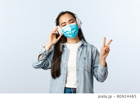 Social distancing lifestyle, covid-19 pandemic and self-isolation leisure concept. Happy smiling asian girl listening music in wireless headphones, wear medical mask and show peace sign Social distancing lifestyle, covid-19 pandemic and self-isolation leisure concept. Happy smiling asian girl listening music in wireless headphones, wear medical mask and show peace sign 80924465