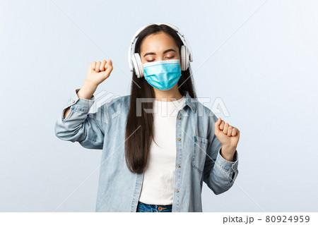 Social distancing lifestyle, covid-19 pandemic and self-isolation leisure concept. Happy upbeat asian girl in medical mask, listening music in wireless headphones and dancing 80924959