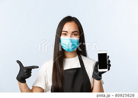 Covid-19 pandemic, coffee shop, small business and preventing virus concept. Attractive asian female employee, cafe worker in medical mask and gloves pointing at mobile phone screen Covid-19 pandemic, coffee shop, small business and preventing virus concept. Attractive asian female employee, cafe worker in medical mask and gloves pointing at mobile phone screen 80925626