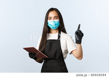 Covid-19, online orders, small coffee shop and preventing virus concept. Smiling friendly asian waitress, female barista in medical mask and gloves point upper right corner, use digital tablet 80926094