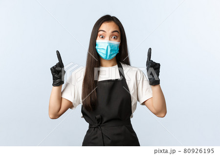 Covid-19 pandemic, social distancing, small business and preventing virus concept. Impressed asian female barista in medical mask and gloves pointing up, stare camera astonished Covid-19 pandemic, social distancing, small business and preventing virus concept. Impressed asian female barista in medical mask and gloves pointing up, stare camera astonished 80926195