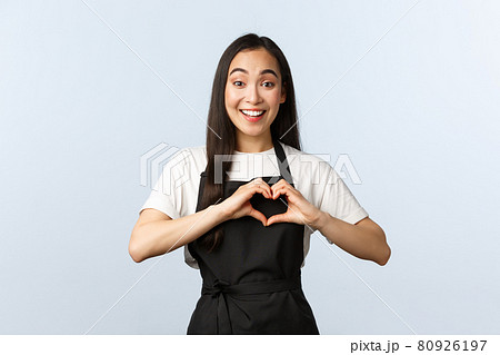 Coffee shop, small business and startup concept. Cute and caring young asian female cafe employee love coworkers or clients, showing heart sign over chest, wearing black apron 80926197