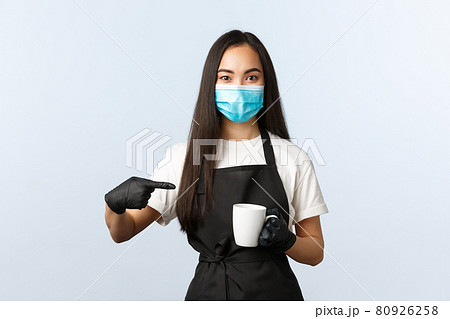 Covid-19, social distancing, small coffee shop business and preventing virus concept. Smiling asian barista in medical mask and gloves recommend try coffee at her cafe, pointing at cup Covid-19, social distancing, small coffee shop business and preventing virus concept. Smiling asian barista in medical mask and gloves recommend try coffee at her cafe, pointing at cup 80926258