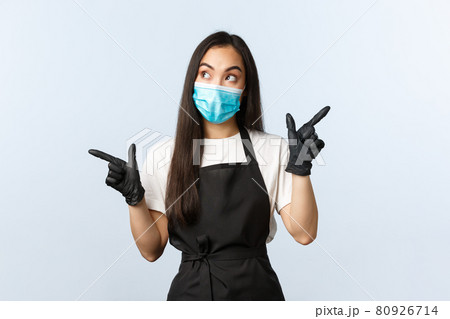 Covid-19, social distancing, small coffee shop business and preventing virus concept. Thoughtful asian barista, cafe owner in medical mask and gloves making choice, pointing sideways 80926714