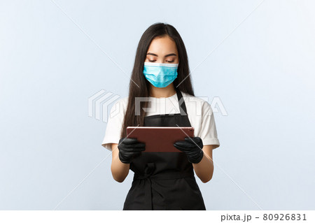 Covid-19, online orders, small coffee shop business and preventing virus concept. Young waitress in medical mask looking at digital tablet as taking order from cafe visitor, barista at work Covid-19, online orders, small coffee shop business and preventing virus concept. Young waitress in medical mask looking at digital tablet as taking order from cafe visitor, barista at work 80926831
