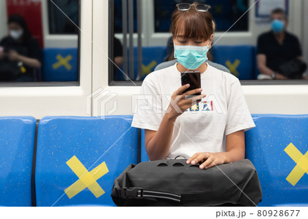 Asian woman with surgical face mask feel tired use smartphone sitting on blue seat in subway, skytrain, carrying backpack, traveling to the city, social distancing, coronavirus, COVID19 80928677