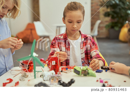 Enthusiastic llittle girl looking concentrated while putting an electronic toy kit together, sitting at the table during STEM class Enthusiastic llittle girl looking concentrated while putting an electronic toy kit together, sitting at the table during STEM class 80931463